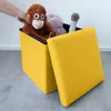 A hand is placing a stuffed monkey into a yellow storage ottoman. The ottoman is open, and other stuffed toys are visible inside. The lid of the ottoman is open and resting against the side. Produktbild-Vorschau