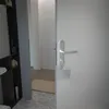 A close-up view of a modern door handle on a white door, with a glimpse of a shelf unit and hallway in the background. Produktbild-Vorschau