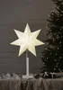 A white star-shaped lamp is displayed on a table with Christmas tree lights in the background. Podgląd zdjęcia produktu
