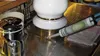 A hand is using a silicone sealant gun to apply sealant around the base of a white and gold decorative item on a metallic ribbed surface. Produktbild-Vorschau