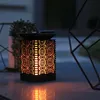 A solar lantern with a flame effect is placed on a surface at dusk, with decorative geometric structures in the background. Podgląd zdjęcia produktu