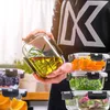 A person is holding a glass food container filled with oil and herbs, with other containers of food stacked nearby. Produktbild-Vorschau