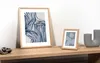 Two framed pictures with abstract blue and white designs are displayed on a wooden shelf, next to a grey sculptural vase. Produktbild-Vorschau