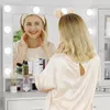 A woman is applying makeup in front of a lighted mirror, with various cosmetic products on a vanity. Voir les images du produit