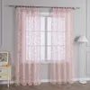 A room interior with pink sheer curtains hanging over a window, a white dresser with a framed picture, and a white floor lamp.