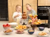 Two young girls are happily using a chocolate fountain with various dipping items. Podgląd zdjęcia produktu