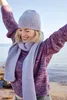 A woman wearing a knitted sweater, hat, and scarf, with her arms raised in the air, standing by the water. Produktbild-Vorschau