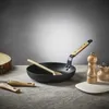 A frying pan with a wooden spoon inside, placed on a textured surface with a wooden log and salt and pepper grinders in the background. Podgląd zdjęcia produktu