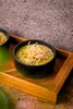A close-up shot of a bowl of green soup topped with crispy fried noodles, served on a wooden tray. Náhľad obrázku produktu