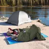 Two people are lying in sleeping bags on a beach next to a lake, with a tent in the background. Náhľad obrázku produktu