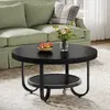 A round black coffee table with a lower shelf is placed in front of a green sofa. A bowl of fruit and a vase of flowers are on the table. Produktbild-Vorschau