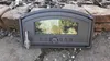A cast iron pizza oven door with a glass window rests on a stone and brick surface, with wood debris in the background. Produktbild-Vorschau
