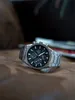 A close-up shot of a silver men's watch with a black dial, resting on a textured brown surface. Produktbild-Vorschau