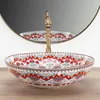 A decorative ceramic basin with a colorful floral pattern sits on a wooden surface, with a brass faucet and a round mirror in the background. Náhled obrázku produktu