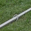 A close-up view of a silver metal object with a curved hook-like attachment, resting on green grass. Produktbild-Vorschau