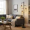 A living room scene featuring a dark gray sectional sofa, a wooden coffee table with books and a mug, a floor lamp with three industrial-style shades, and a window with beige curtains. Produktbild-Vorschau