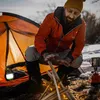 A man is warming his hands over a campfire next to an orange tent, with a camping lantern visible inside the tent. Produktbild-Vorschau
