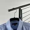 A close-up view of a light blue collared shirt hanging on a black hanger, which is suspended from a black metal rack. The background is plain white. Produktbild-Vorschau
