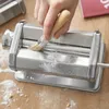 A hand is using a brush to clean a pasta maker on a wooden table with scattered flour. Produktbild-Vorschau