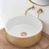 A close-up shot of a decorative washbasin with gold accents, set against a white wall and a wooden countertop. Náhled obrázku produktu