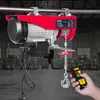 A red electric hoist is mounted on a metal bar in a warehouse setting, with a hand holding a remote control. Náhled obrázku produktu