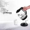 A hand is pouring water from a glass electric kettle into a white mug. The kettle has a black lid and base, and the mug is white. The Lehmann Home logo is visible on the left side of the image. Shadows of leaves are cast on the background. Produktbild-Vorschau