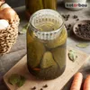 A close-up shot of a glass jar filled with pickles and a white plastic fermentation weight on top, set against a rustic wooden background with various ingredients like onions, carrots, and peppercorns scattered around. Anteprima dell’immagine prodotto