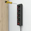 A black power strip with four outlets and individual switches is mounted on a white wall, with a yellow and gray screwdriver about to screw in a nail at the top. Produktbild-Vorschau