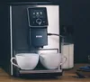 A coffee machine is shown making coffee into two cups, with a milk container next to it. Produktbild-Vorschau