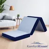 A foldable mattress is set up in a living room next to a sofa and a small side table with a plant. Voir les images du produit