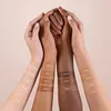 Four hands of different skin tones are held together, with foundation swatches applied to the forearms. Produktbild-Vorschau