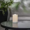 A white LED candle sits on a glass-topped table next to a large potted plant. Produktbild-Vorschau