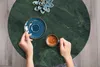A top-down view of hands holding a cup of coffee and a spoon on a green marble table. Náhľad obrázku produktu