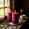 Two large red pillar candles burning on a wooden table next to a window, with a copper teapot and a cup of tea in the background. Anteprima dell’immagine prodotto