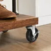 Close-up view of a wooden shelf with a black caster wheel and a brown shoe on top. A white cabinet is visible in the background. Náhled obrázku produktu