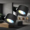 Two black wall-mounted spotlight fixtures are shown in a modern interior setting. The spotlights are angled, with their lights casting a warm yellow and cool blue glow. Anteprima dell’immagine prodotto