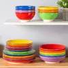 Colorful ceramic bowls and plates are neatly stacked on white shelves and a wooden surface. Náhled obrázku produktu