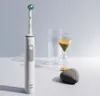 An electric toothbrush, hourglass, glass, and stone are arranged on a white surface against a light gray background. Produktbild-Vorschau