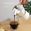 Coffee being poured from a moka pot into a glass cup on a wooden table. Produktbild-Vorschau
