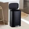 A black pedal trash can with its lid open, placed next to a kitchen counter and cabinet. A round woven rug is visible on the floor. Produktbild-Vorschau