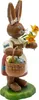 Ein niedlicher Osterhase aus Holz steht auf einer grünen Basis und hält einen Korb voller bunter Ostereier in der einen Hand während er in der anderen Hand einen Strauß gelber Blumen trägt und ein charmantes Lächeln zeigt Produktbild-Vorschau