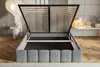 A bed frame with the mattress support mechanism lifted, revealing the storage space underneath. The bed is in a bedroom setting with a side table and lamp. Náhled obrázku produktu