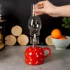 A hand is placing the glass chimney onto a red and white polka dot oil lamp. In the background are wooden discs and a bowl of oranges. Náhled obrázku produktu