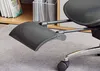 A close-up of a black office chair's footrest and part of the seat, resting on a gray carpet. Books are visible on a shelf in the background. Náhled obrázku produktu