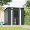 A dark gray metal garden shed with open doors, revealing a ladder and rake inside. It is situated on a wooden deck next to a wooden fence and a green hedge, with grass in the foreground. Náhled obrázku produktu