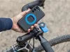 A person is holding a smartphone with a Quad Lock case attached, demonstrating how it connects to a bike mount on the handlebars. Náhled obrázku produktu