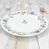 A white oval plate with a maritime design is placed on a light-colored wooden surface, partially covered by a burlap cloth. A wine glass is tipped over in the background, and a small glass votive with a candle is to the left. Seashells are scattered in the Podgląd zdjęcia produktu