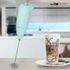 A mint green Clatronic milk frother stands next to a glass of coffee on a table, with a kitchen in the background. Produktbild-Vorschau