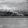 A close-up, low-angle view of the edge of a rug with a textured, abstract pattern in black, gray, and white, resting on a wooden floor. Produktbild-Vorschau