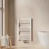 A white towel radiator is mounted on a light beige wall in a bathroom. To the left, a wooden stool with a folded towel and some small containers is placed near a window. To the right, a portion of a white bathtub is visible. Produktbild-Vorschau